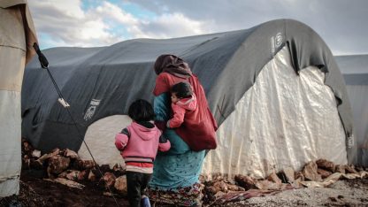 Mulher carregando criança no colo em frente a tendas de um campo de refugiados em terreno rochoso. — Foto: Ahmed akacha