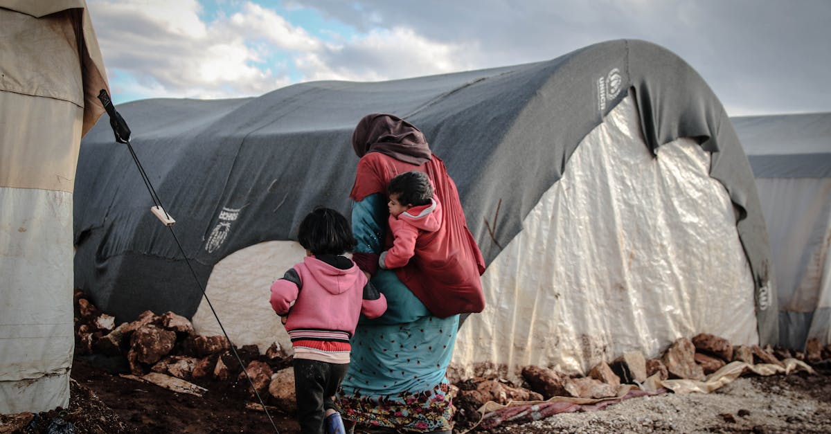 Mulher carregando criança no colo em frente a tendas de um campo de refugiados em terreno rochoso. — Foto: Ahmed akacha