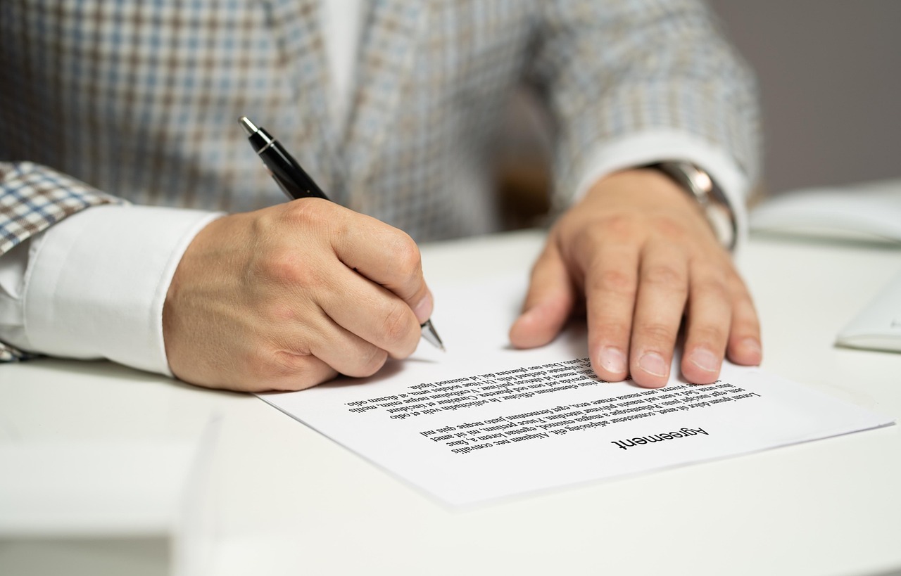 Close das mãos de um homem de terno assinando um documento de contrato sobre uma mesa branca. — foto: tumisu