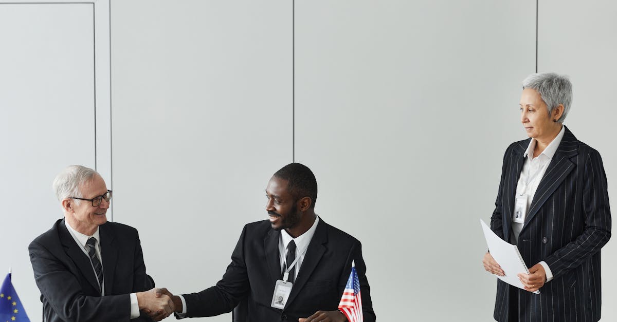Dois executivos em trajes formais apertando as mãos em uma mesa com bandeiras internacionais e uma mulher ao lado. — Foto: Werner Pfennig