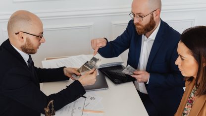 Dois advogados de terno e uma cliente analisando fotos e documentos sobre uma mesa branca em um escritório. — Foto: www.kaboompics.com
