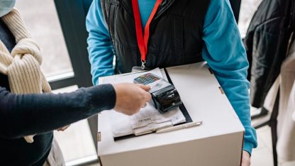 Entregador segurando caixa de papelão enquanto cliente realiza pagamento em máquina de cartão. — Foto: Yan Krukau
