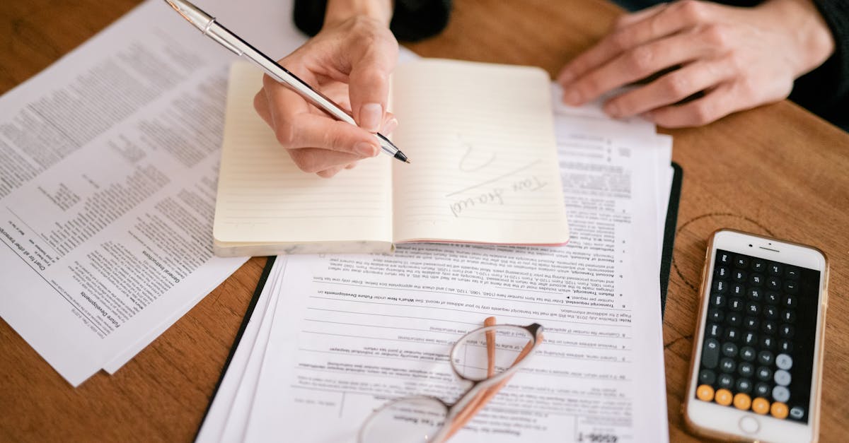 Mãos de uma pessoa escrevendo em caderno sobre mesa com formulários de impostos, óculos e celular com calculadora. — Foto: Leeloo The First