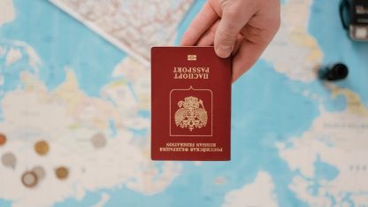 Mão segurando um passaporte vermelho sobre um mapa múndi com moedas espalhadas ao fundo. — Foto: Tima Miroshnichenko