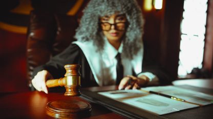 Juíza com peruca clássica e toga segurando um martelo de madeira sobre mesa com documentos em um tribunal. — Foto: khezez  | خزاز