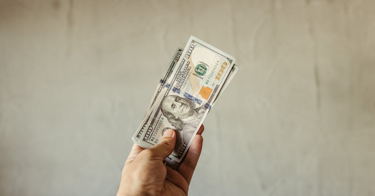 Mão segurando notas de cem dólares americanos contra um fundo neutro. — Foto: Jonathan Borba