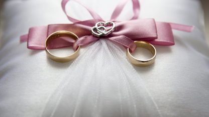 Duas alianças de ouro sobre uma almofada branca com laço rosa e detalhes em formato de coração. — Foto: Pixabay