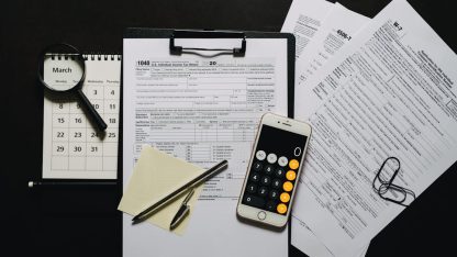 Documentos fiscais em uma prancheta ao lado de um celular com calculadora, lupa, caneta e calendário sobre mesa preta. — Foto: Leeloo The First