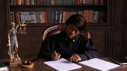 Juíza negra escrevendo em documentos em uma mesa de madeira com a estátua da justiça e livros ao fundo. — Foto: KATRIN  BOLOVTSOVA