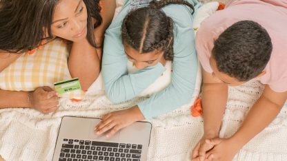 Mulher e duas crianças deitadas usando um notebook e segurando um cartão bancário sobre uma manta branca. — Foto: Kindel Media