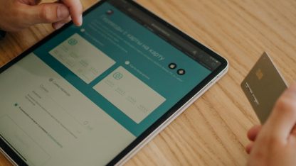 Pessoa segurando cartão de crédito e utilizando tablet para realizar pagamento online em mesa de madeira. — Foto: Tima Miroshnichenko