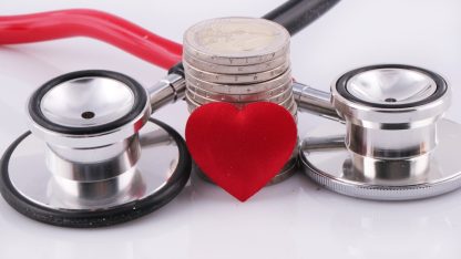 Um estetoscópio médico ao lado de uma pilha de moedas e um pequeno coração vermelho sobre fundo branco. — Foto: Marek Studzinski