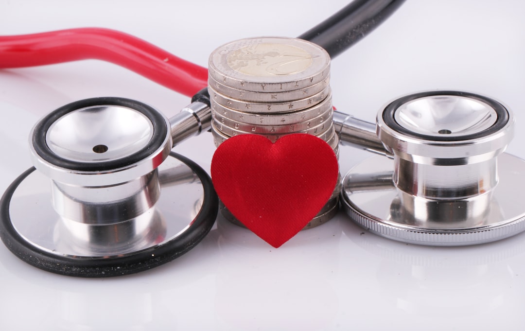 Um estetoscópio médico ao lado de uma pilha de moedas e um pequeno coração vermelho sobre fundo branco. — Foto: Marek Studzinski