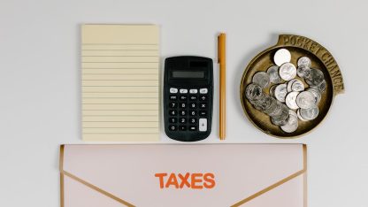 Calculadora, bloco de notas, moedas e uma pasta com a palavra Taxes organizada sobre uma mesa branca. — Foto: Tara Winstead