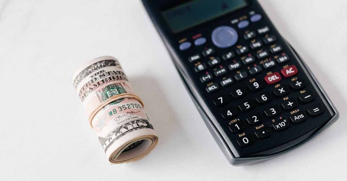 Um rolo de cédulas de dinheiro ao lado de uma calculadora científica sobre uma superfície branca. — Foto: www.kaboompics.com