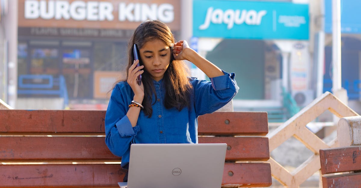 Mulher jovem sentada em um banco de praça usando notebook e falando ao celular, com lojas ao fundo. — Foto: Shantanu Kumar