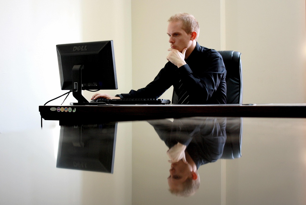 Homem em traje social concentrado trabalhando em um computador em escritório moderno com reflexo na mesa. — Foto: Pexels