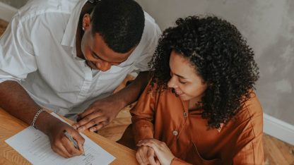Homem e mulher negros sorridentes assinando um documento sobre uma mesa de madeira. — Foto: olia danilevich
