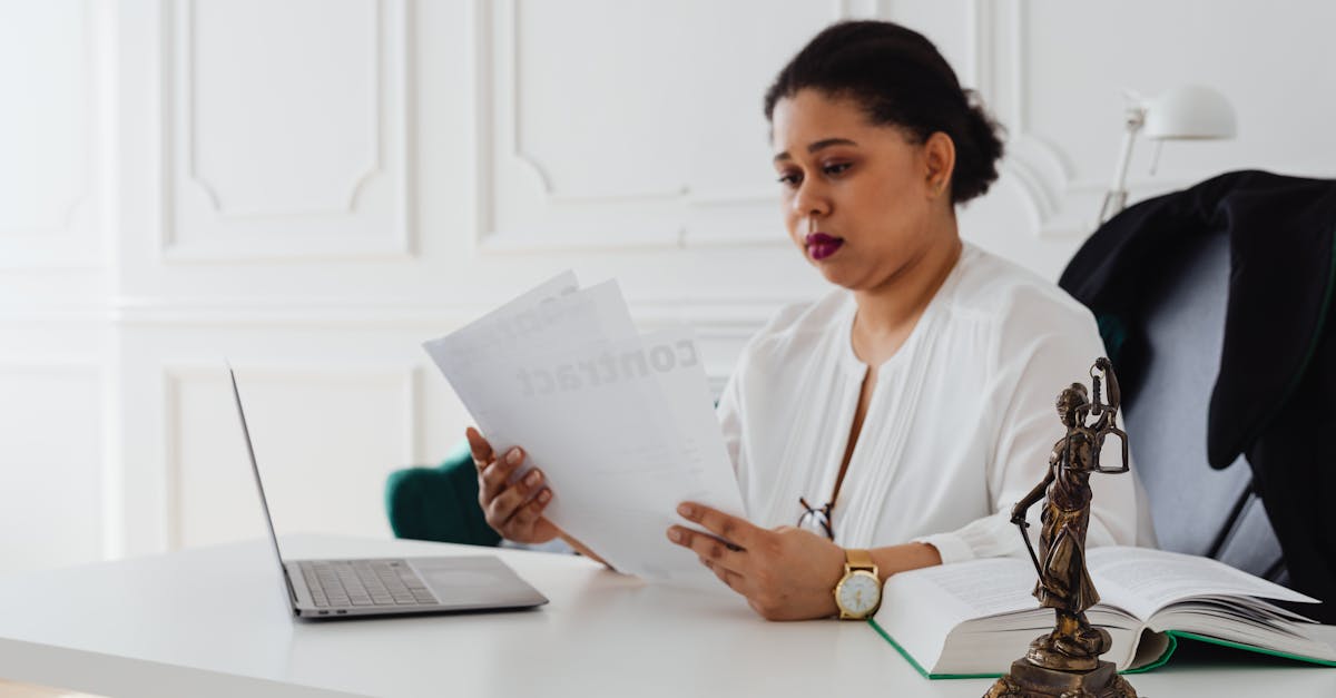 Advogada analisando documentos em escritório com notebook, livro jurídico e estátua da justiça sobre a mesa. — Foto: www.kaboompics.com