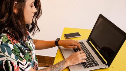 Mulher jovem com tatuagem no braço trabalhando em um notebook sobre uma mesa amarela. — Foto: Ândrea Ferreira