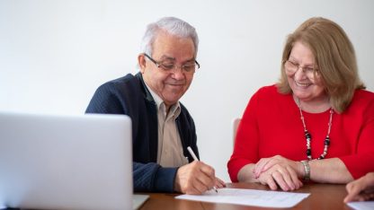 Casal de idosos sorridentes assinando papéis em uma mesa com notebook em ambiente de escritório. — Foto: Kampus Production