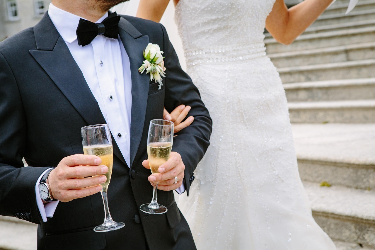 Noivo de smoking e noiva de vestido branco segurando taças de champanhe em uma escadaria. — Foto: Pexels