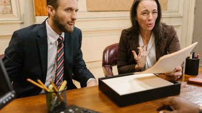 Dois advogados em trajes formais discutem documentos em uma mesa de escritório durante atendimento a cliente. — Foto: RDNE Stock project