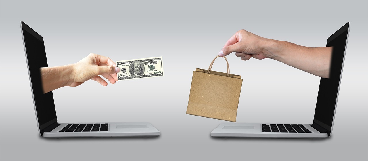 Mãos trocando dinheiro e sacola entre laptops, simbolizando compra online. — Foto: Mediamodifier