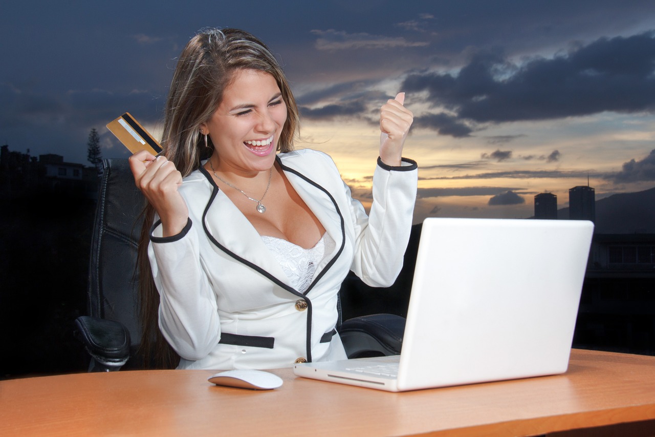 Mulher sorridente segurando cartão de crédito em frente a um notebook, representando uma compra online. — Foto: DigitalMarketingAgency
