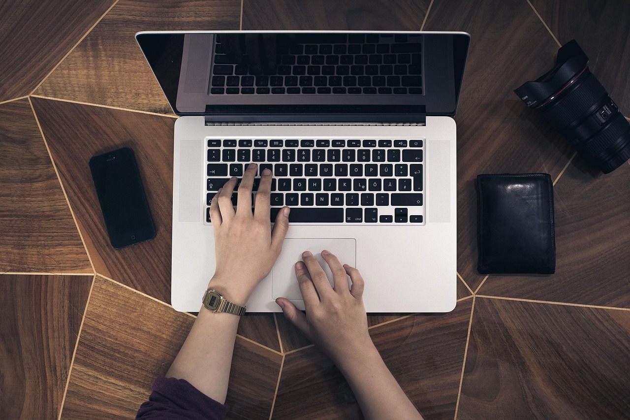 Pessoa digitando em um laptop com itens pessoais ao lado, em uma mesa de madeira. — Foto: Pexels