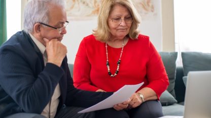 Casal de idosos sentados em um sofá analisando papéis e documentos com um notebook à frente. — Foto: Kampus Production