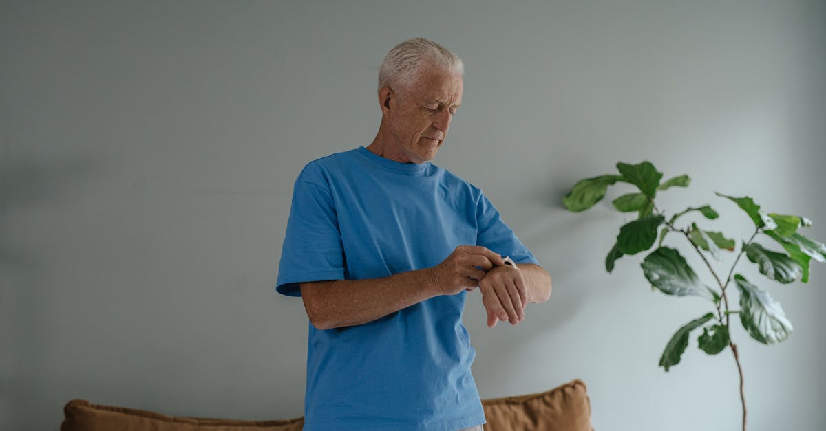 Homem idoso de camiseta azul olhando para o relógio de pulso em um ambiente interno com uma planta ao fundo. — Foto: SHVETS production