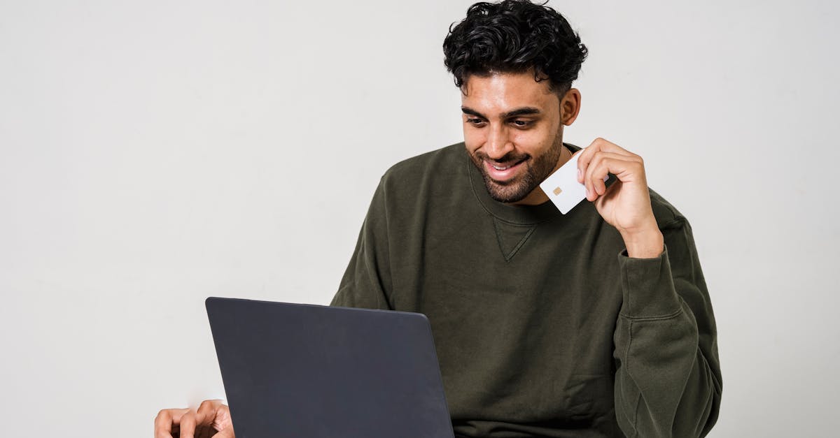 Homem sorridente segurando um cartão de crédito branco enquanto utiliza um notebook em fundo claro. — Foto: Sora Shimazaki