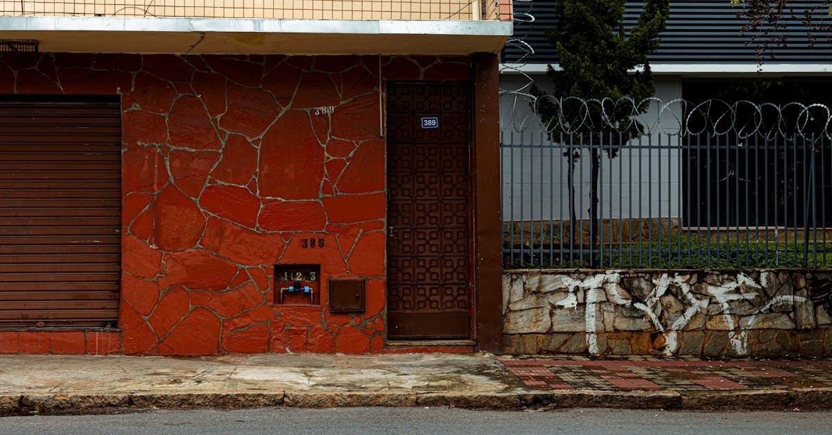 Fachada de casa com parede de pedra vermelha, porta de metal e grade com cerca elétrica em rua urbana. — Foto: Laura Villela Beauty Designer | Brasil