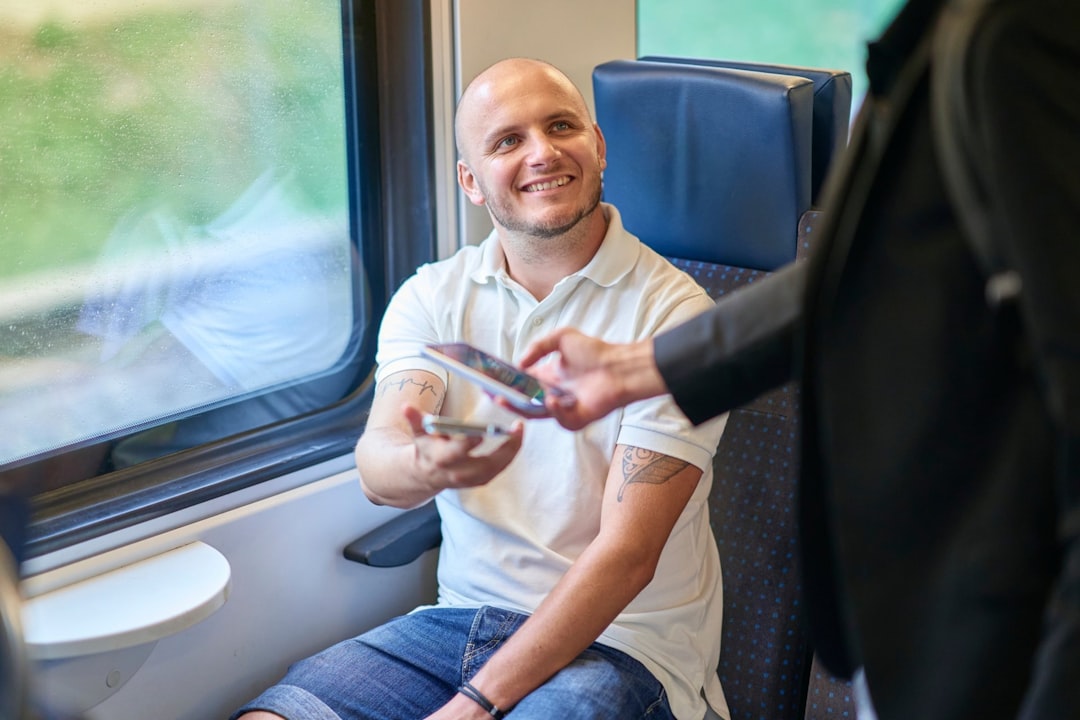 Homem sorridente sentado em um trem entregando um bilhete ou cartão para outra pessoa. — Foto: FAIRTIQ AG