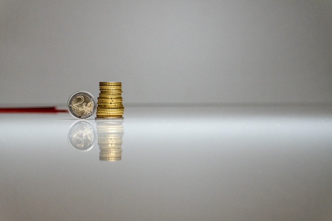 Pilha de moedas douradas e uma moeda de dois euros sobre uma superfície branca reflexiva. — Foto: John Vid