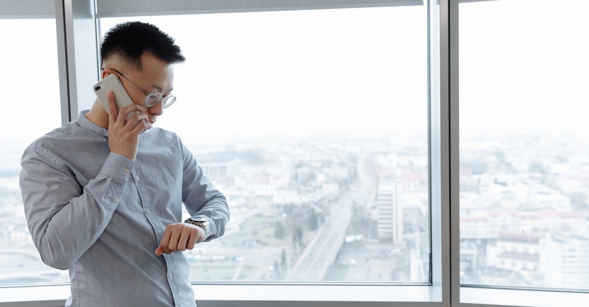 Homem de camisa social falando ao celular e olhando para o relógio em um escritório com vista para a cidade. — Foto: Thirdman