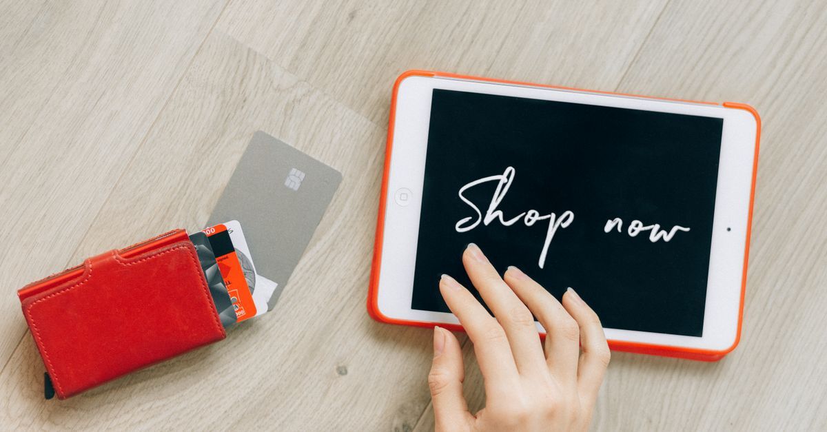 Mão usando tablet com mensagem 'shop now' e carteira com cartões de crédito sobre uma superfície de madeira. — foto: nataliya vaitkevich