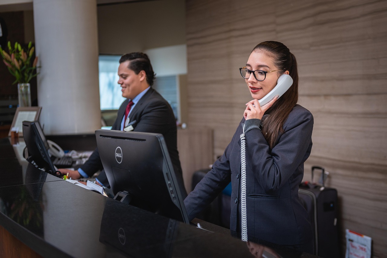 Atendente de hotel em conversa telefônica, com outro funcionário no fundo, em ambiente de trabalho. — foto: rodrigo_salomonhc