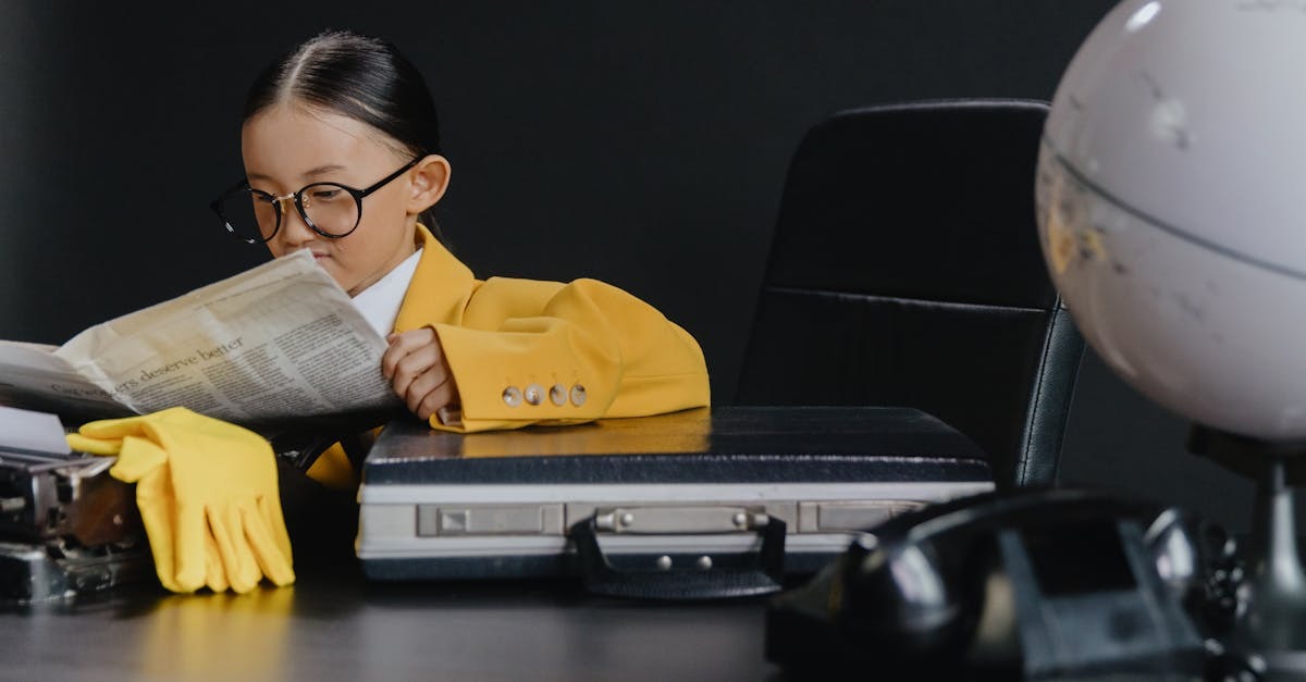 Menina usando óculos e casaco amarelo lendo um jornal em frente a uma mala e telefone. — Foto: cottonbro studio