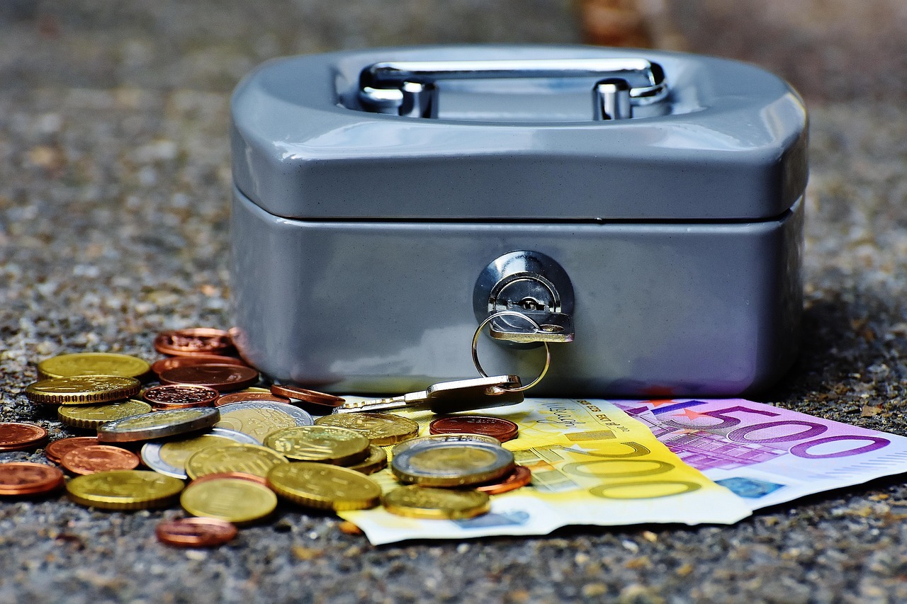 Caixa de metal com moedas e cédulas espalhadas no chão.