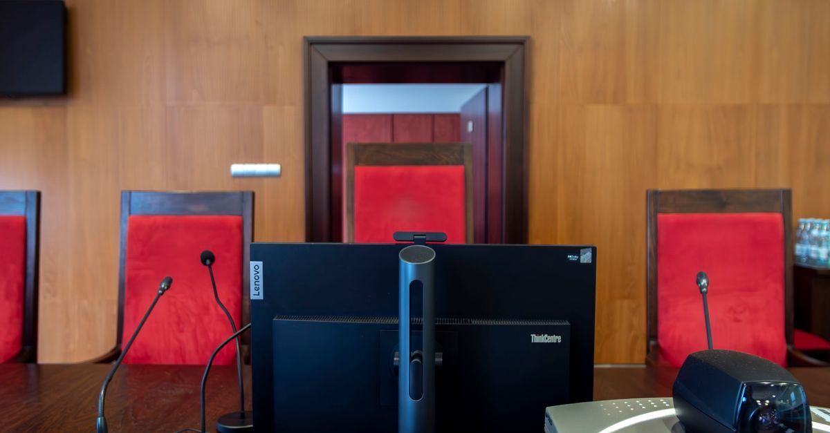 Sala de audiência com cadeiras vermelhas e microfones, ambiente de um processo judicial.