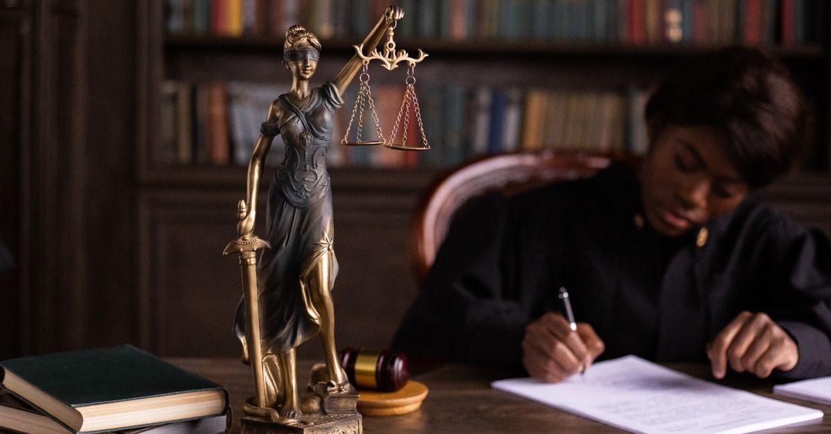 Pessoa escrevendo em um documento em frente a uma estátua de justiça e livros, sugerindo um contexto legal.