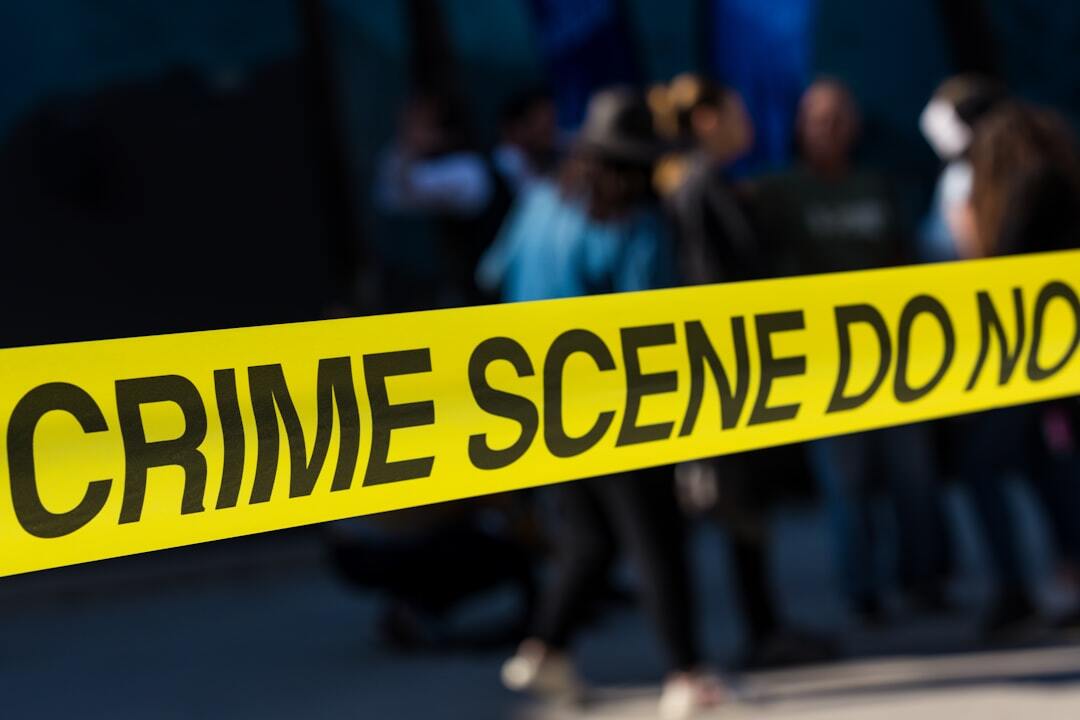 Fita amarela de isolamento com a inscrição crime scene em destaque com pessoas desfocadas ao fundo.