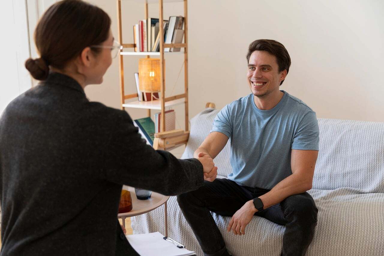 Duas pessoas em uma sala, uma dando uma mão no ombro da outra, sugerindo apoio ou conversa sobre bem-estar.