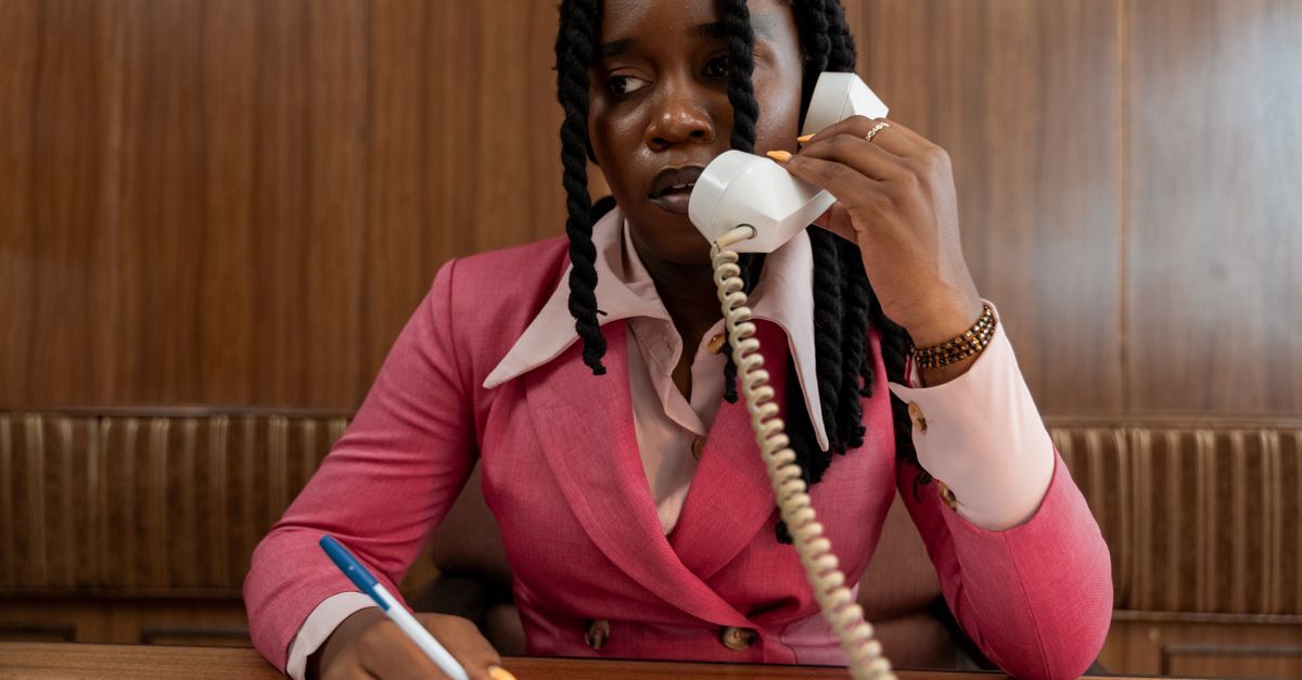 Cobranca indevida inline 2 103823 1787218585 Mulher em blazer rosa conversando ao telefone, com caneta na mão, em ambiente de escritório.