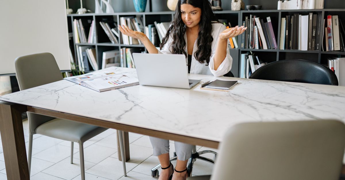 Mulher em frente a um laptop em uma mesa de trabalho, com livros no fundo, sugerindo um ambiente de trabalho ou estudo.