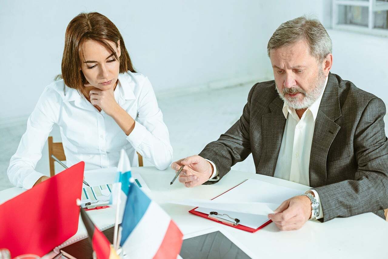 Duas pessoas em uma mesa discutindo documentos, uma com caneta em mãos, em ambiente de trabalho.