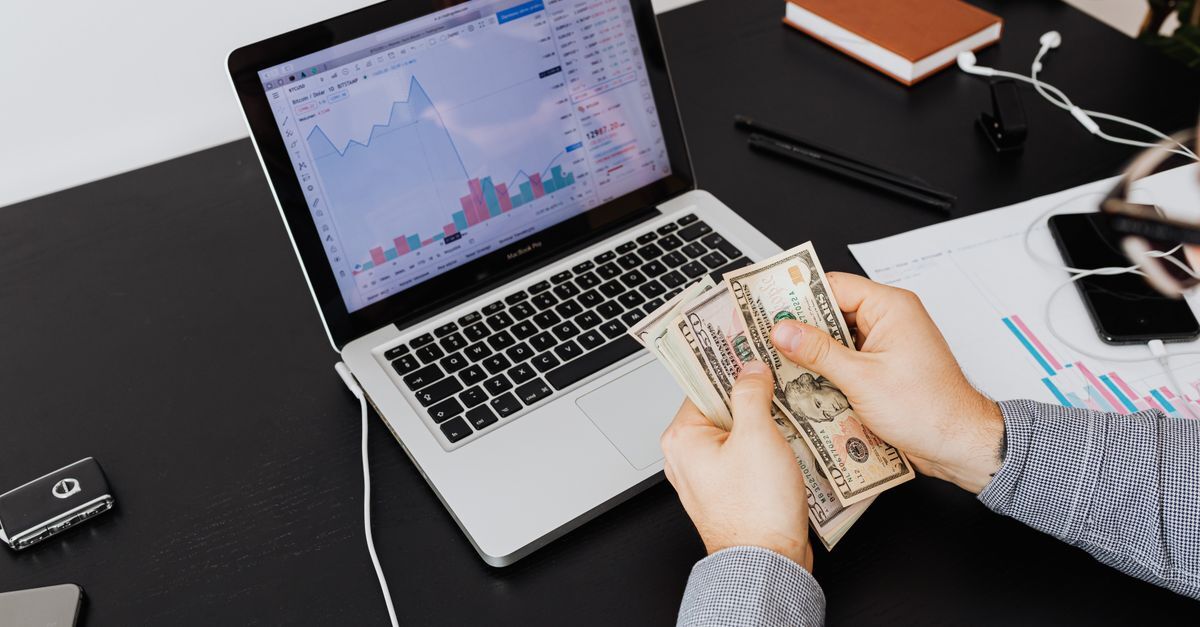 Pessoa segurando dinheiro em frente a um laptop com gráficos financeiros.