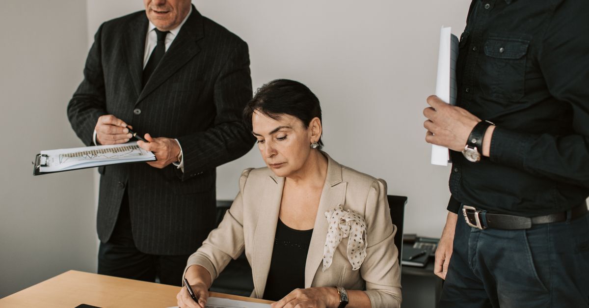 Mulher assinando documentos em uma mesa com dois homens observando.